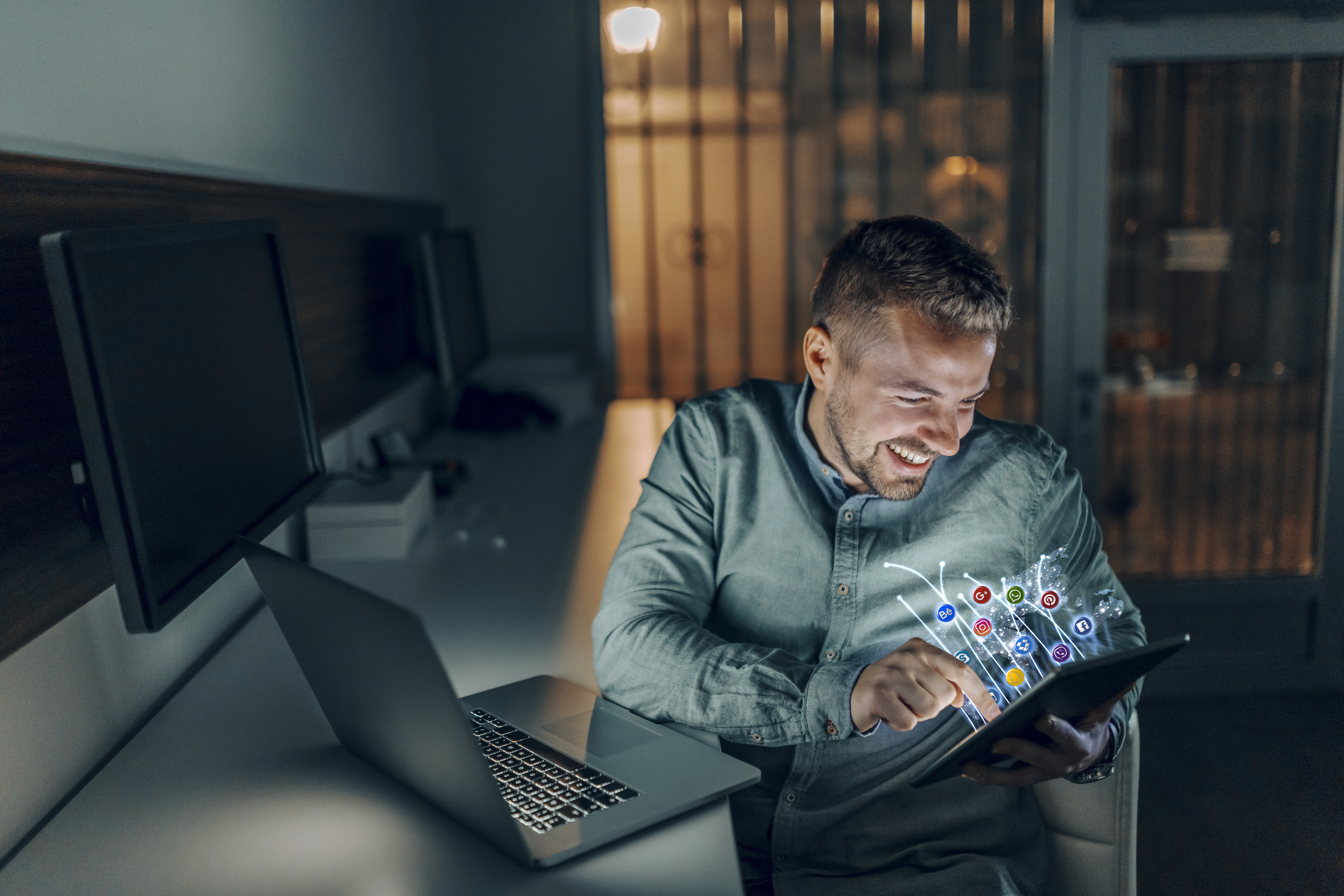 Handsome Caucasian bearded freelancer with toothy smile sitting in office late at night and using tablet for surfing the internet. Social media concept.